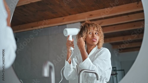 happy woman dressed white bathrobe looking at the mirror smiling dries blond curly hair with a hairdryer slow motion photo