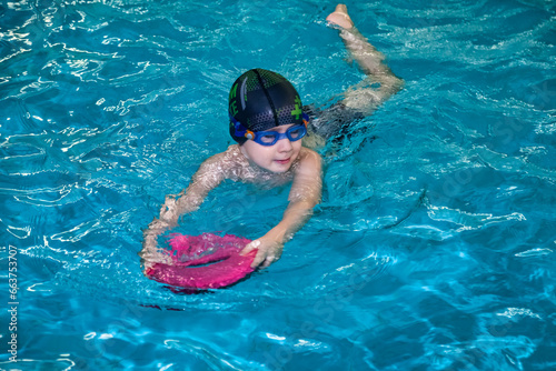 Child boy swimmer in swimming suit training with float in swim pool, sport learn. Kid five year old in swim cap and swimming goggles exercising. Workout learning children concept. Copy ad text space