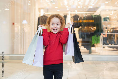 Happy child girl in red sweater with shopping bags in cloth store. Black Friday concept