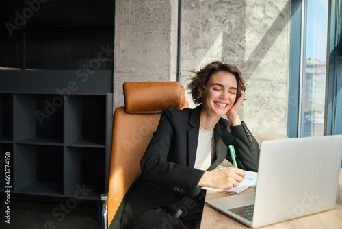 Portrait of woman listening to webinar in her office, joing online meeting, writing down, making notes in documents, sitting near laptop photo