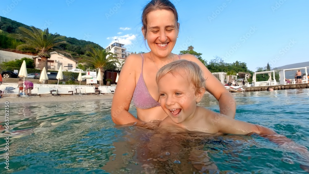 Happy laughing toddler boy learns swimming in sea with mother. Family holiday, summertime, fun vacation on beach