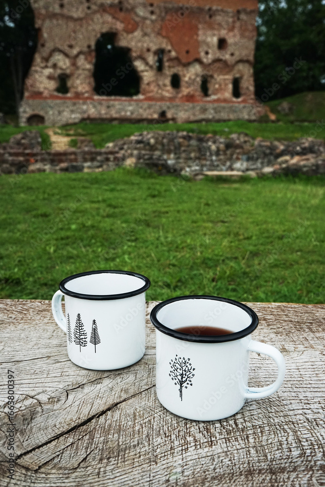 Obraz premium Two white metal mugs on a wooden bench against the background of ruins and forest