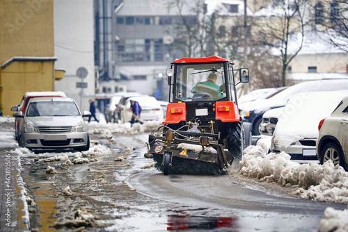 Tractor with rotating brush sweep road from snow in residential area. Snow removal tractor cleans parking lot after blizzard. Utility vehicle sweeping asphalt road. Snowplough work, clean pavement