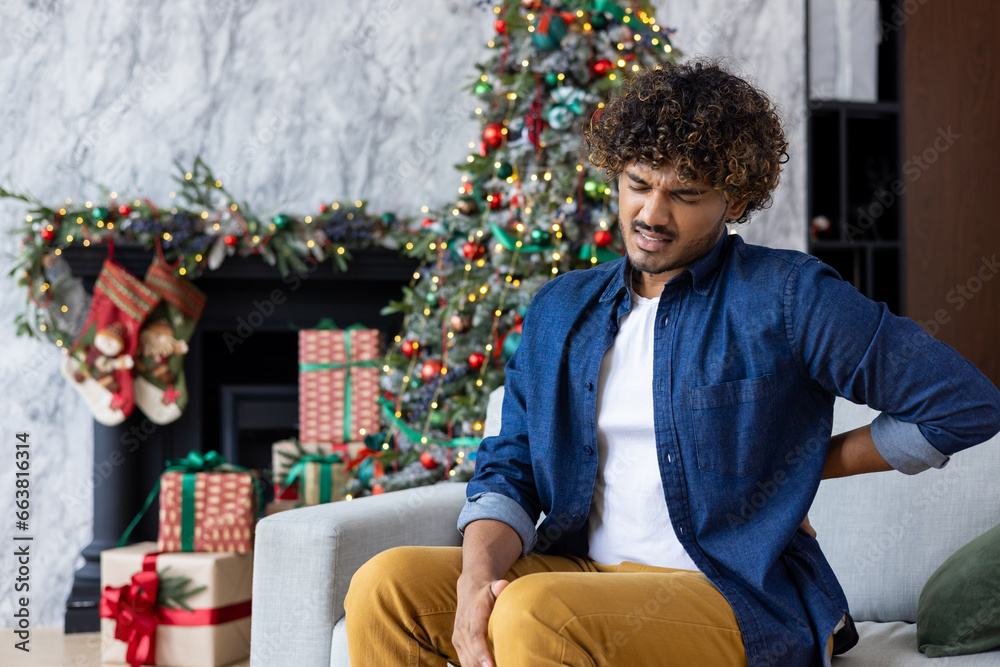 A sick man on Christmas has severe back pain, massages muscle his handб sitting on sofa in living room. Hispanic celebrating New Year and Christmas holidays on a winter day near tree with gifts. A sick man on Christmas has severe back pain, massages muscle his handб sitting on sofa in living room. Hispanic celebrating New Year and Christmas holidays on a winter day near tree with gifts.