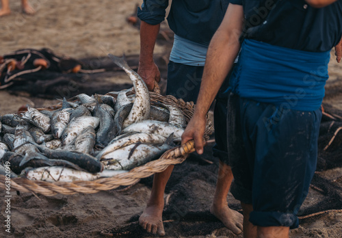 Unrecognizable barefoot fishermen waking with basketful of fishes on beach photo