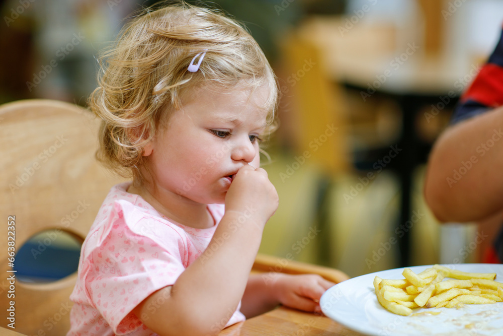 Adorable toddler girl eating healthy vegetables and unhealthy french fries potatoes. Cute happy baby child taking food from dish at daycare or nursery canteen.
