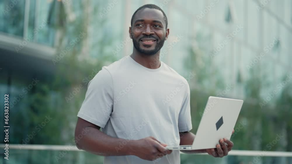 African american man working on laptop looking aside smiling standing on street near modern building. Portrait bearded positive guy in city downtown. Lunch break ending work office lifestyle concept. African american man working on laptop looking aside smiling standing on street near modern building. Portrait bearded positive guy in city downtown. Lunch break ending work office lifestyle concept.