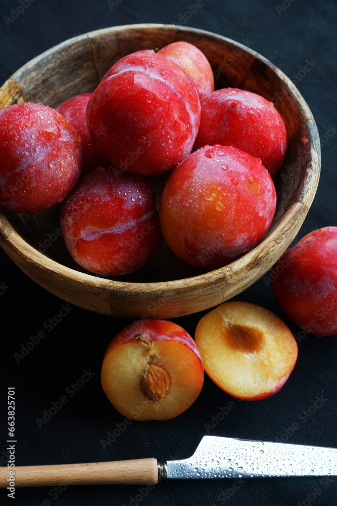 Ripe plums in a bowl next to sliced plums and a knife on a dark tablecloth Ripe plums in a bowl next to sliced plums and a knife on a dark tablecloth