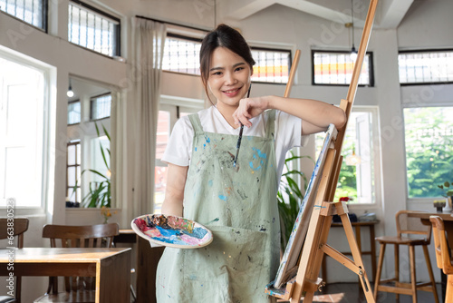 Portrait of young woman painting holding a palette and paintbrush in front of a canvas in a painting studio.