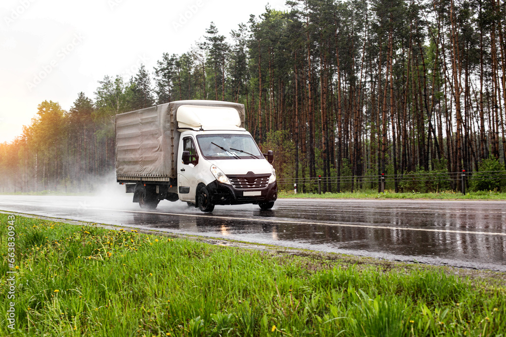 A small truck transports cargo against the backdrop of a forest on a wet road in the rain, industry