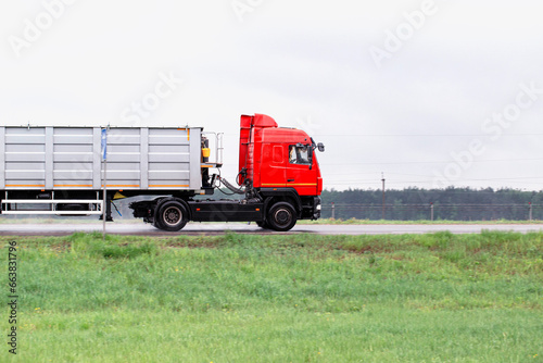 A red truck with a semi-trailer transports bulk cargo along the road in cloudy weather. Side view. Copy space for text, industry
