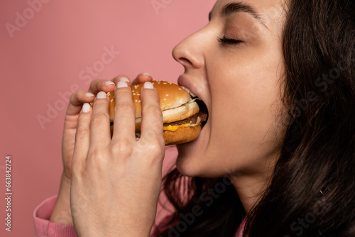 Closeup of a dark-haired woman eating an appetizing cheeseburger in front of the camera on the pink background. Hunger and favorite fast food concept