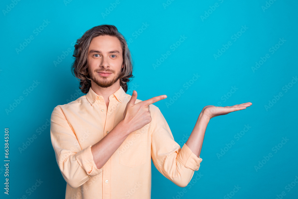Photo of cheerful pleasant man with long hairstyle dressed yellow shirt directing at empty space on palm isolated on blue color background Photo of cheerful pleasant man with long hairstyle dressed yellow shirt directing at empty space on palm isolated on blue color background
