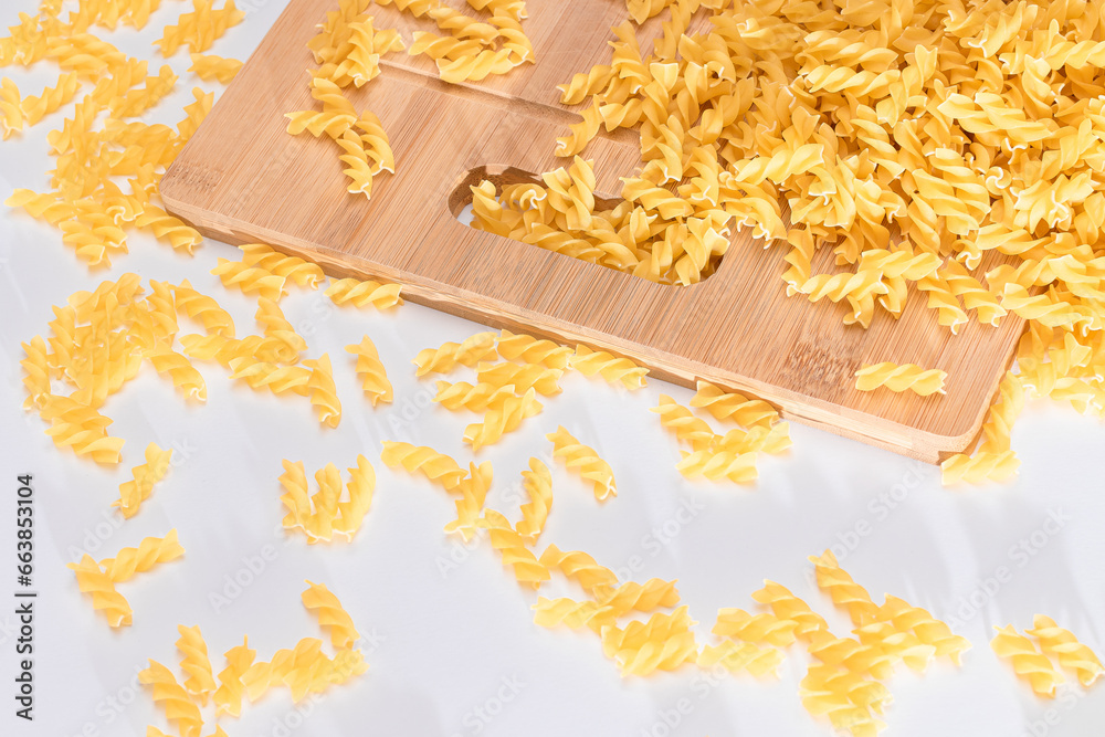 Obraz premium Uncooked Fusilli Pasta Scattered on Wooden Board and White Table. Raw and Dry Macaroni. Fat and Unhealthy Food