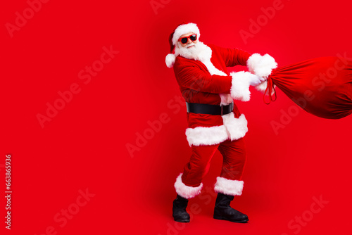 Full size photo of funky grandfather wear santa costume throwing bag with presents to empty space isolated on red color background.