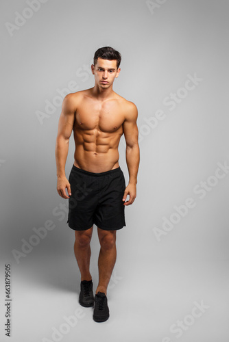 Full length image of a man with an athletic and fit body, posing in the studio with a bare torso, showing six abs pack, looking at camera, grey background.