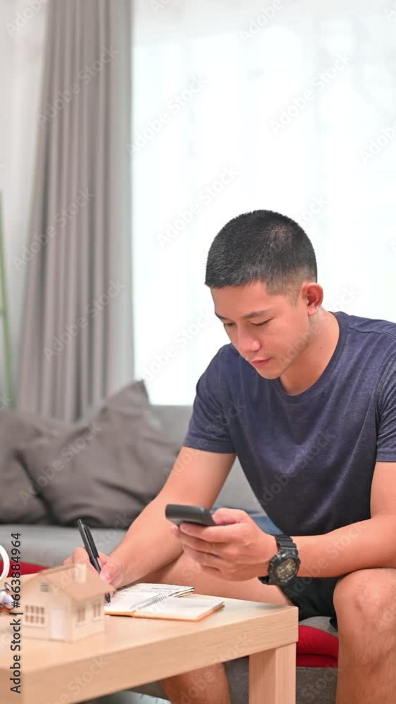 Handsome millennial man using smart phone for making online payments, managing expenses finances in living room Handsome millennial man using smart phone for making online payments, managing expenses finances in living room