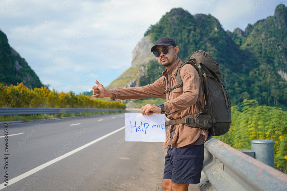 Asian man holding a poster: Please help for hitchhiking. Male tourist worried about traffic jam on empty rural road