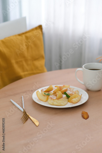 Autumn home interior. Breakfast of cottage cheese with peach, ceramic vase with autumn leaves, candle, glass of coffee on round wooden table..