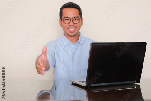 A man sitting in front of a laptop offering handshakes with happy expression photo