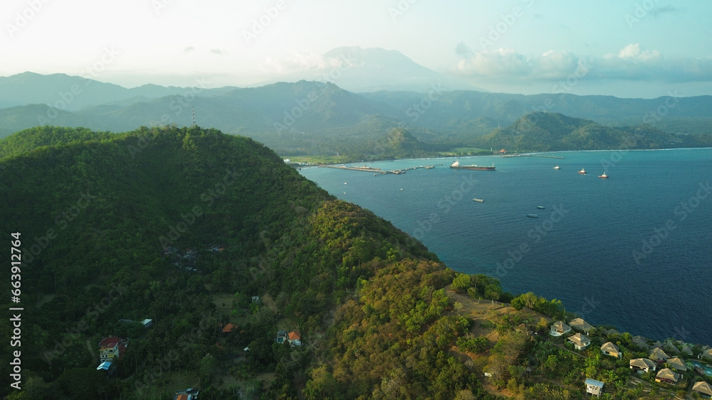 Aerial view of a hill illuminated by golden sunset light at Padang Bai port on the tropical island of Bali. View of sunken ships and the hotel where tourists relax. Aerial view of a hill illuminated by golden sunset light at Padang Bai port on the tropical island of Bali. View of sunken ships and the hotel where tourists relax.