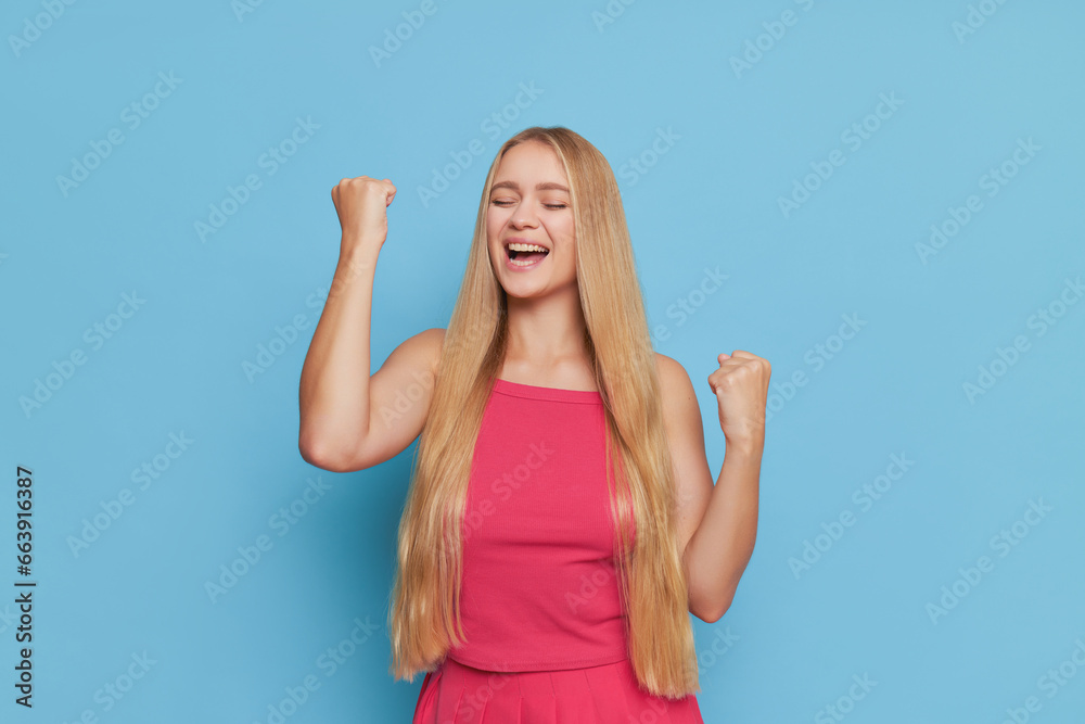 Pretty girl in pink top with long fair hair havig fun over background holding her fists up with joy, wonderful life concept, copy space