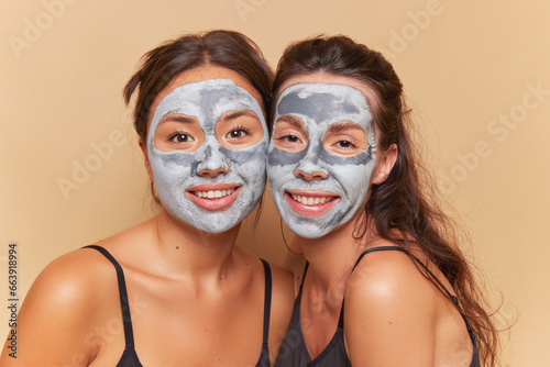Two women with cosmetic clay masks on their faces having relaxing spa procedure with smiles, healthcare product concept, copy space