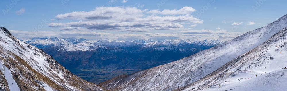 Breathtaking view of The Remarkables ski resort, Coronet Peak, showcasing pristine snow-covered slopes, dotted with skiers against a backdrop of majestic mountains and cloudy skies.