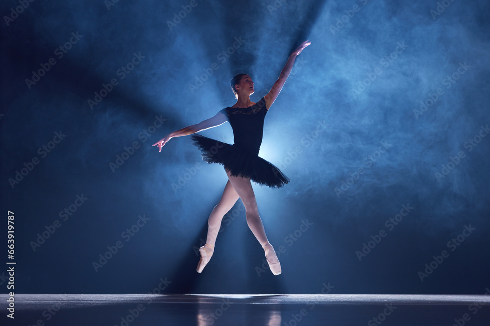 Elegant, artistic, tender ballerina, young woman dancing in tutu on stage over spotlight. Blue light. Concept of classical dance, art and grace, beauty, choreography, inspiration