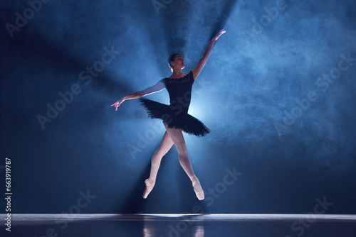 Elegant, artistic, tender ballerina, young woman dancing in tutu on stage over spotlight. Blue light. Concept of classical dance, art and grace, beauty, choreography, inspiration