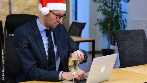 Middle age Caucasian man in black suit and Santa hat is sitting in the office and working on laptop. Businessman is looking around, taking off Santa hat and resting a glass of sparkling wine. photo