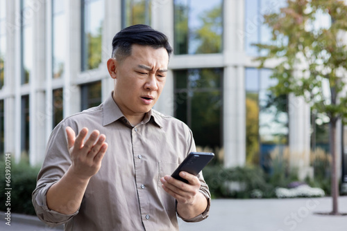 Worried senior asian male office worker businessman standing outside and using mobile phone. Disappointingly looks at the screen photo