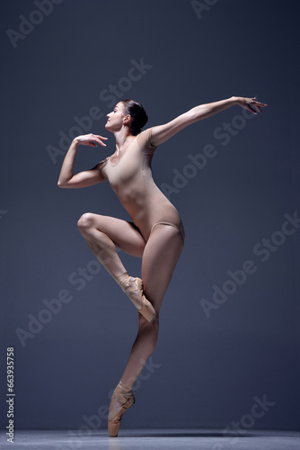 Elegant, attractive woman, professional ballerina in beige bodysuit, standing in pointe against blue studio background. Concept of classical dance, art and grace, beauty, choreography, inspiration