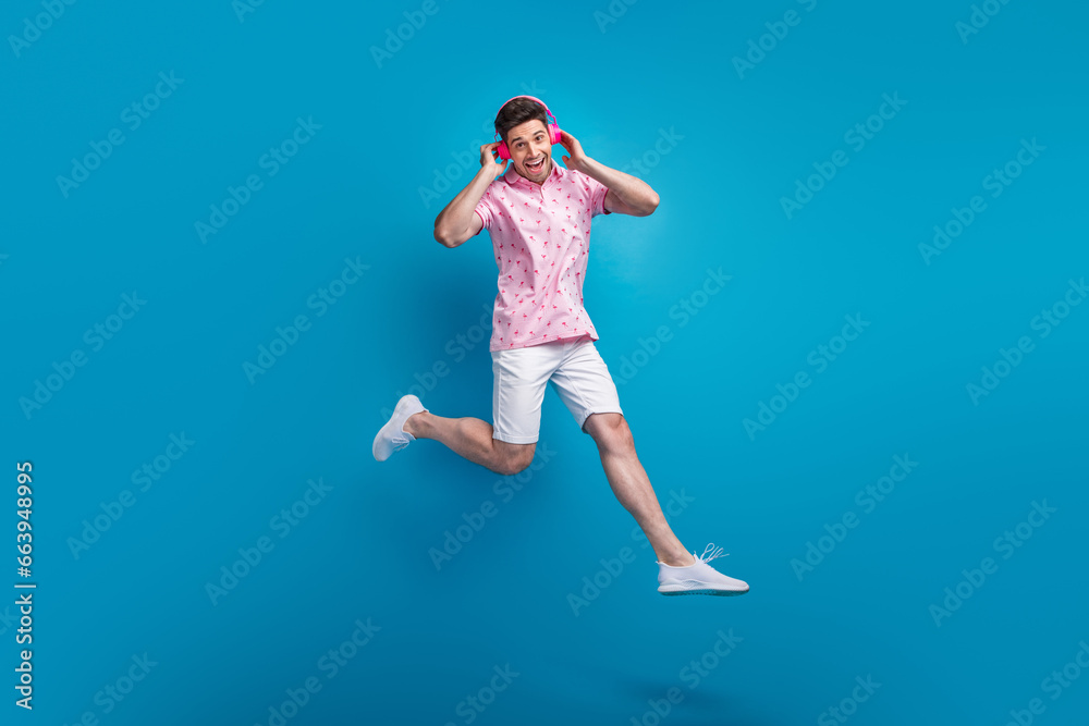 Full length portrait of carefree overjoyed person jumping hands touch headphones isolated on blue color background
