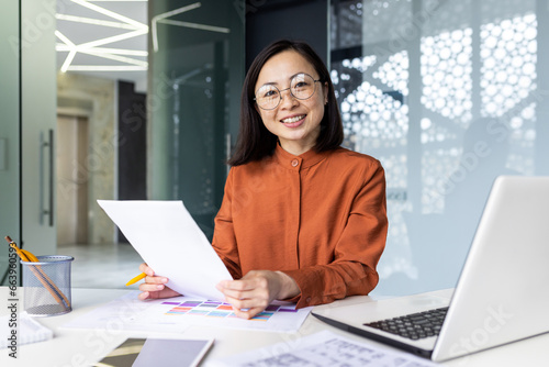 Successful Asian woman doing paperwork inside the office, businesswoman holding documents, reports and papers in her hands, smiling and looking at the camera, portrait of a satisfied financial woman. © Liubomir