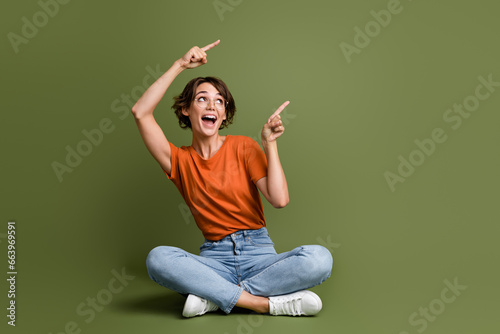 Full length photo of impressed girl dressed t-shirt sit on floor look directing at promo empty space isolated on green color background