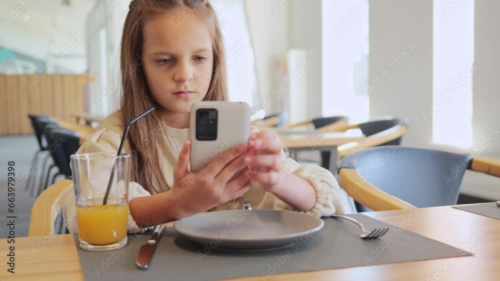 Beautiful little girl wearing casual clothing resting in cafe enjoying drinking fresh juice and using cell phone surfing internet via smartphone. Beautiful little girl wearing casual clothing resting in cafe enjoying drinking fresh juice and using cell phone surfing internet via smartphone.