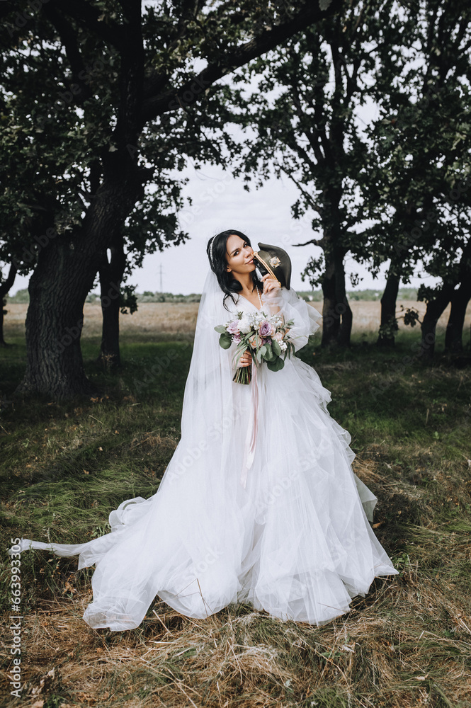 A beautiful military bride with a cap in her hands in a white dress stands in a park in nature. Wedding photography, portrait.