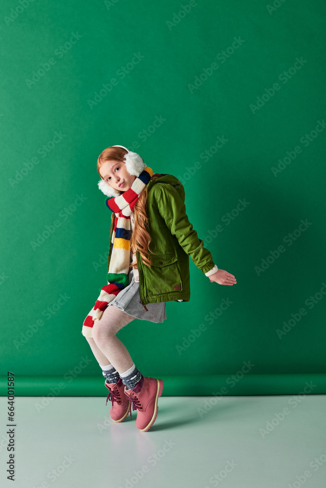stylish preteen girl in ear muffs, striped scarf and winter outfit posing on turquoise backdrop