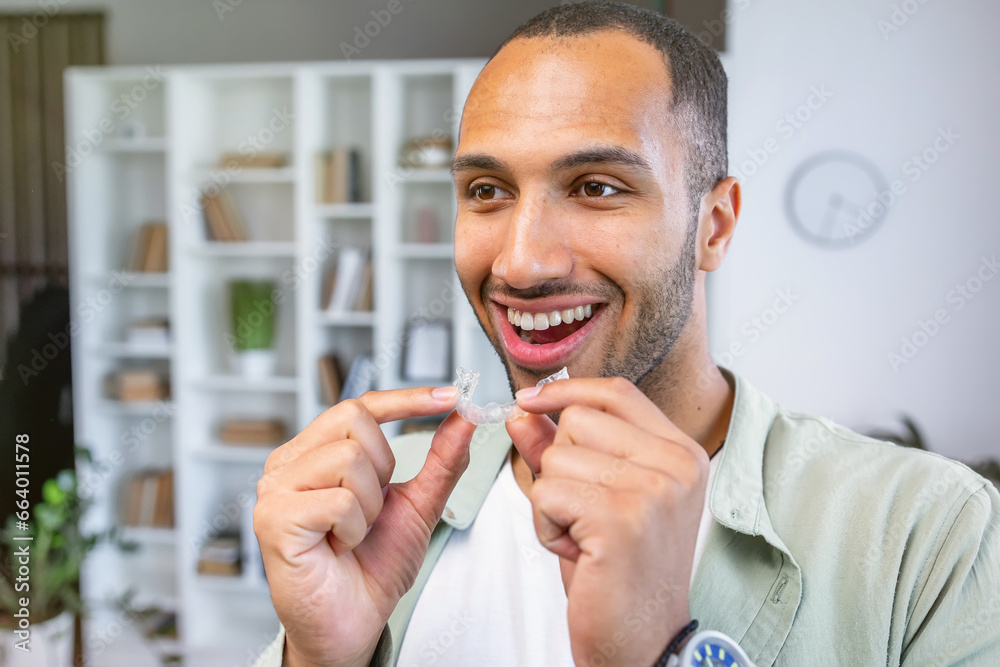 Young man holding dental aligner at home with a happy face standing and smiling with a confident smile showing teeth - obrazy, fototapety, plakaty 
