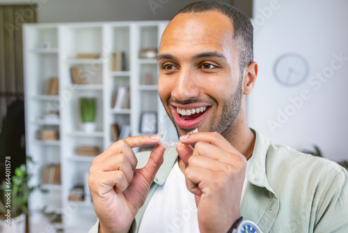 Young man holding dental aligner at home with a happy face standing and smiling with a confident smile showing teeth