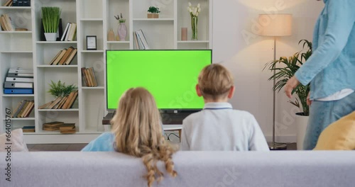 Rear View. Young man and woman son and daughter sitting on couch and watching green screen chroma key TV with fun with smiles. Beautiful family is watching TV on chroma key at home in living room.