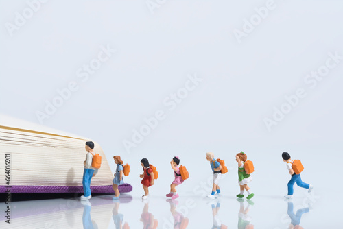 Schoolchildren with schoolbags stand in a row in front of an open book, miniature figures scene, white background, copy space