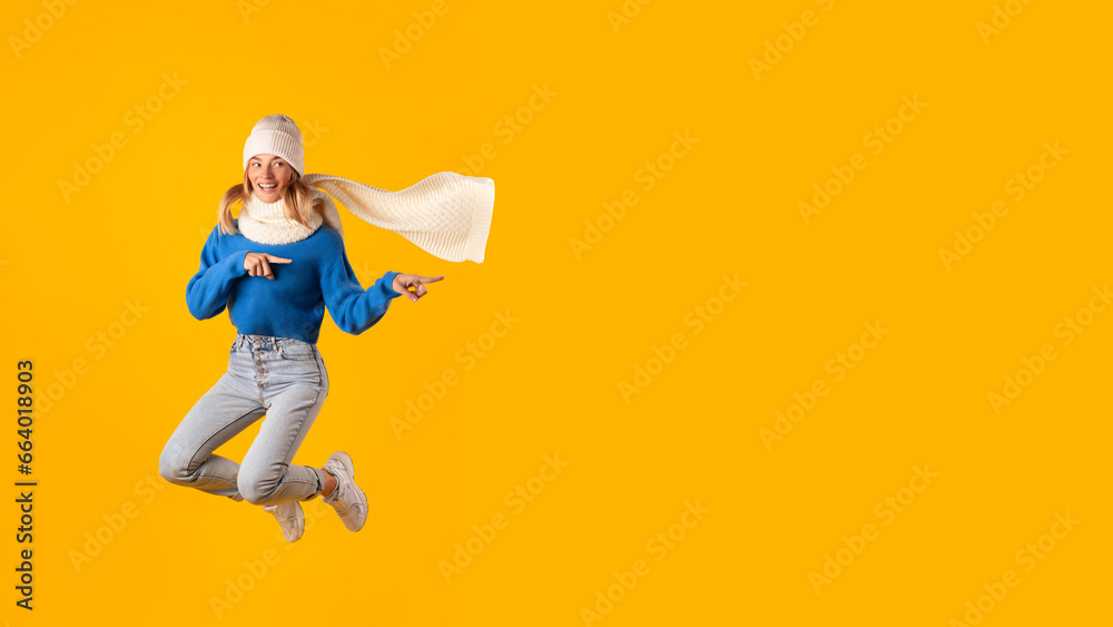 Excited caucasian woman in knitted hat jumping and pointing aside at free space on yellow studio background Excited caucasian woman in knitted hat jumping and pointing aside at free space on yellow studio background