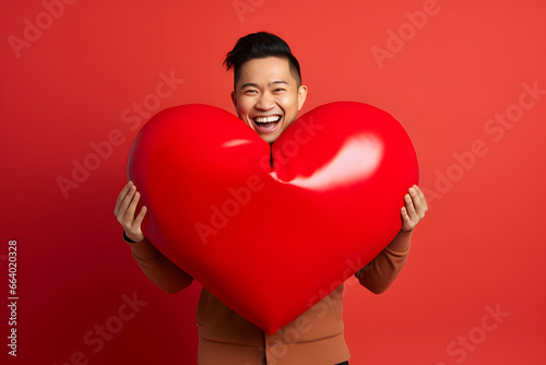 smiling Asian man holding big red heart on red background. Not based on any actual person or scene © lucky pics