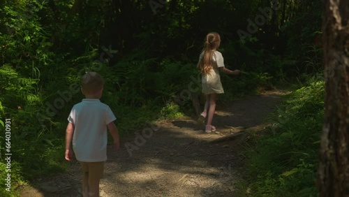 Couple of children moves along shaded forest path backside view. Preschooler boy and schoolgirl rest actively on vacation slow motion. Leisure in nature photo