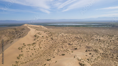 A large sand dune in the middle of the steppe. Drone view of a huge pile of sand. Tourists are walking  enjoying the view. A river runs in the distance and grass grows. The sky with white clouds