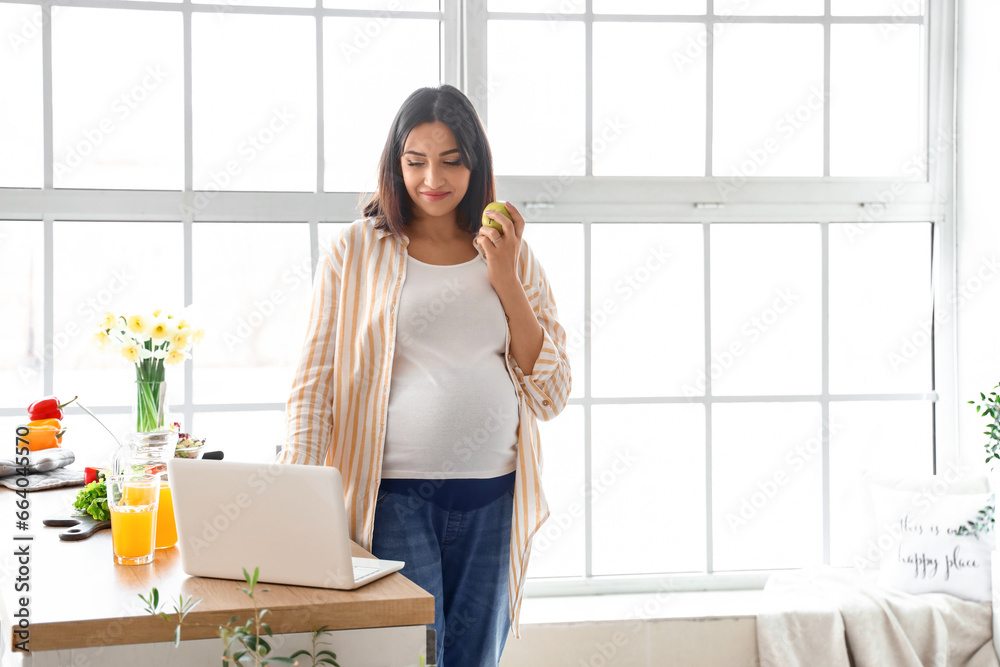 Young pregnant woman with apple using laptop in kitchen Young pregnant woman with apple using laptop in kitchen