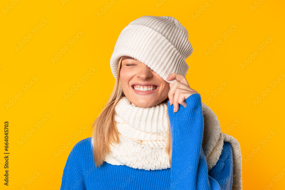 Happy woman playfully pulling knitted hat over one eye and sharing a broad smile over yellow studio background