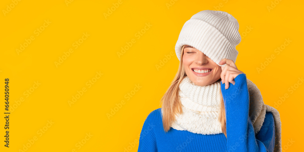 Bright winter offer. Carefree lady covering one eye with white knitted hat, yellow background, panorama with copy space Bright winter offer. Carefree lady covering one eye with white knitted hat, yellow background, panorama with copy space